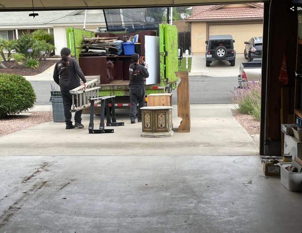 Two Junk MD professionals loading a ladder and furniture into a green truck for a residential garage cleanout service.