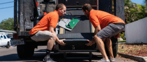 Two Junk MD professionals in orange shirts loading a large treadmill with exposed electronic components into a hauling truck.