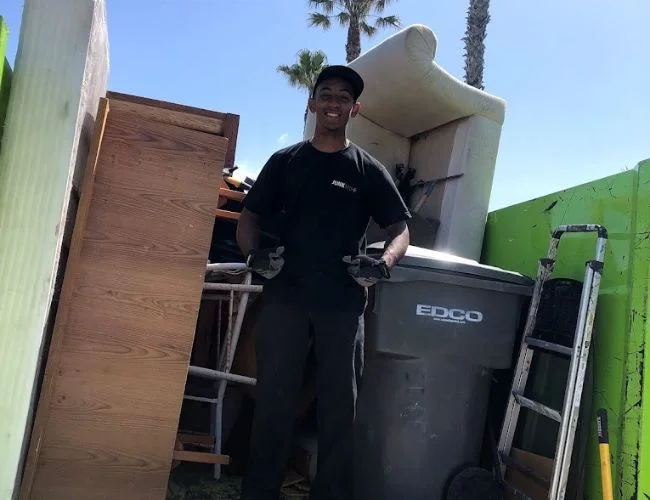 Smiling Junk MD professional in a black uniform standing next to old furniture, a ladder, and an EDCO trash bin in a green hauling truck.