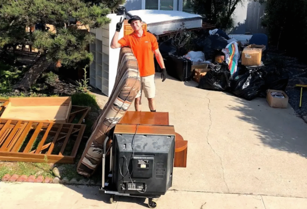 Professional junk removal worker in an orange shirt posing next to a mattress and old CRT television on a residential driveway for disposal.