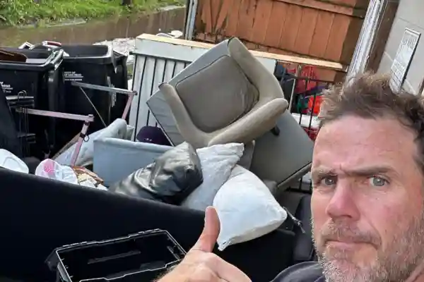 Junk removal professional giving a thumbs up in front of a truck loaded with household debris, old furniture, and trash bags.